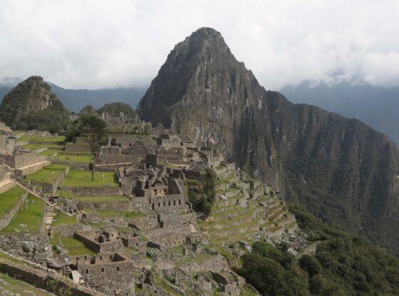 Machu Picchu archaeological site with terraced fields and ancient stone structures, beneath a cloud-covered mountain.